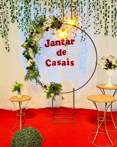 JANTAR DE CASAIS: arco de folhagens com placa, tapete vermelho, mesas altas. Festa realizada no Espaço Comemora em 2024