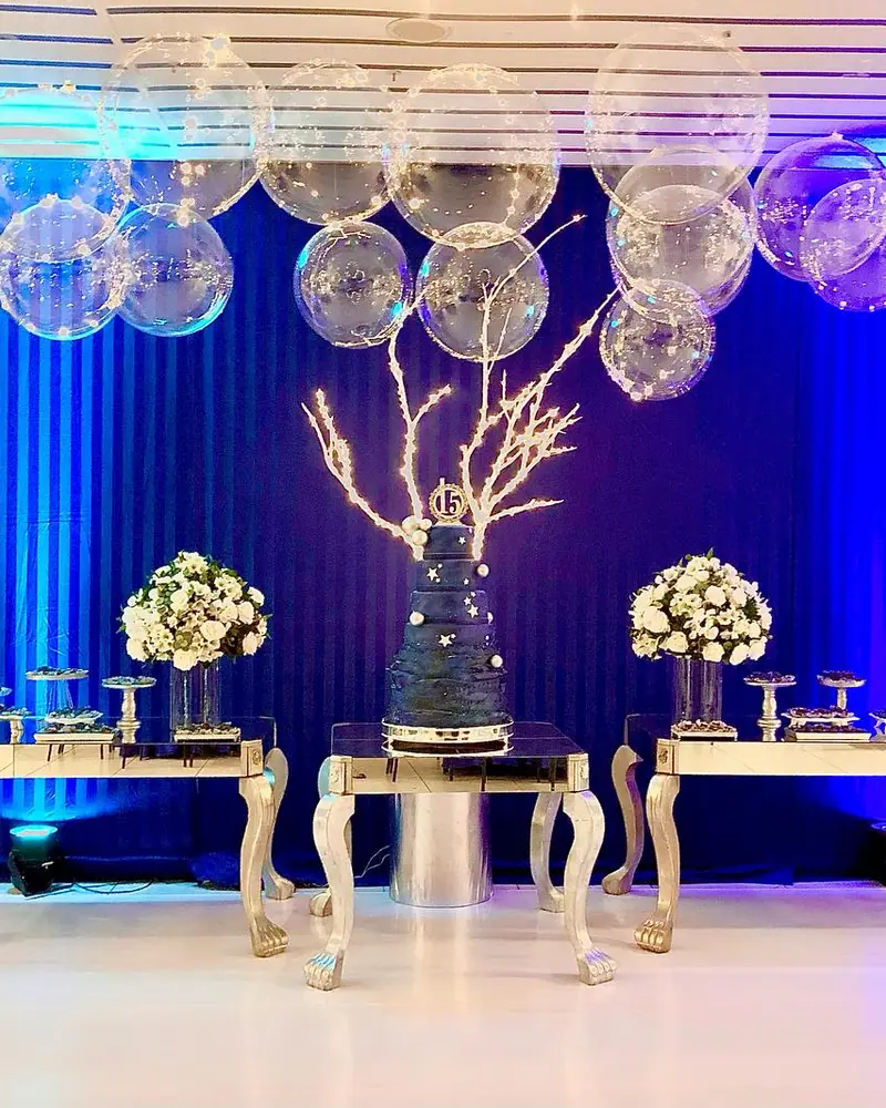 Festa de 15 anos: cortina azul iluminada, balões transparentes com luzinhas e mesa prata