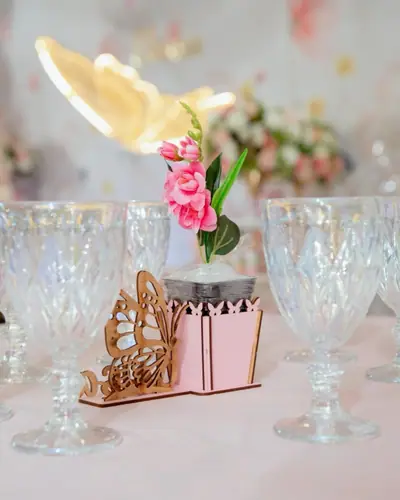 detalhe de mesa: borboleta de MDF rosa, flores, tacas furta-cor; mae marcada @dayaneferreira08. Festa realizada no Espaço Comemora em 2026